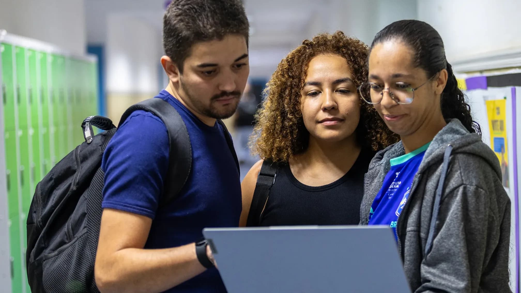 Três estudantes analisam juntos a tela de um notebook em corredor escolar