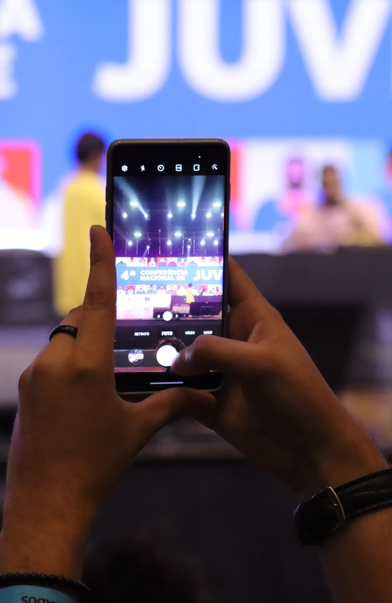 Mãos de jovem erguem o celular para registrar o palco da 4ª Conferência Nacional da Juventude